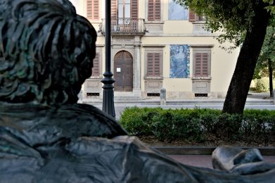 Statua di Giacomo Puccini vista da dietro, con la palazzina regia sullo sfondo